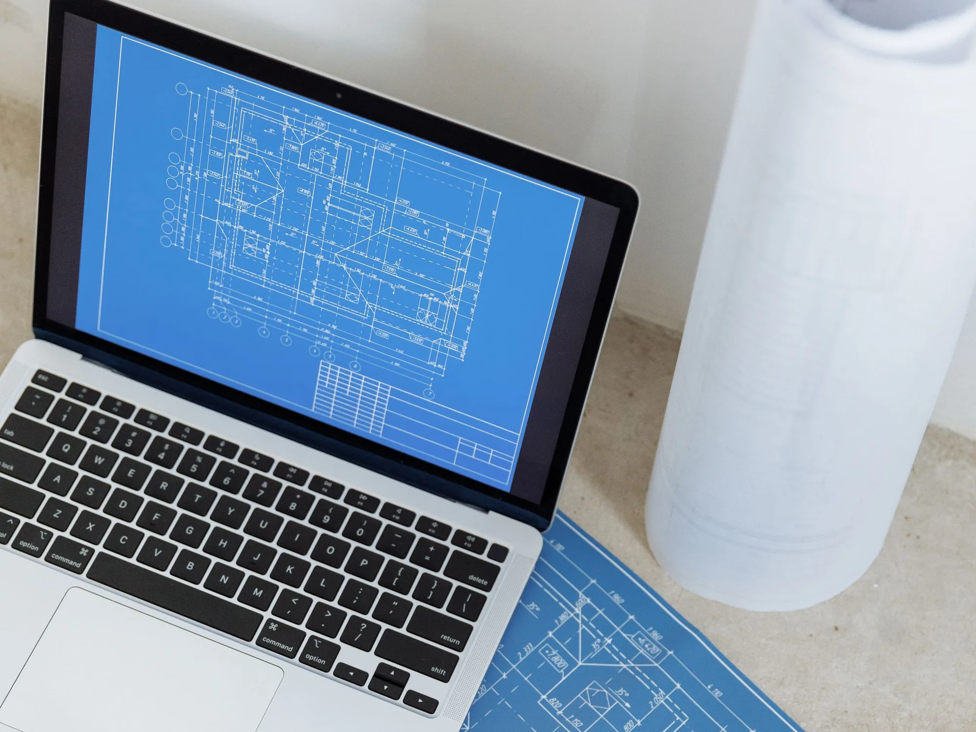Laptop showing a floor plan in blueprint blue, beside a printed blueprint and a roll of plan sets