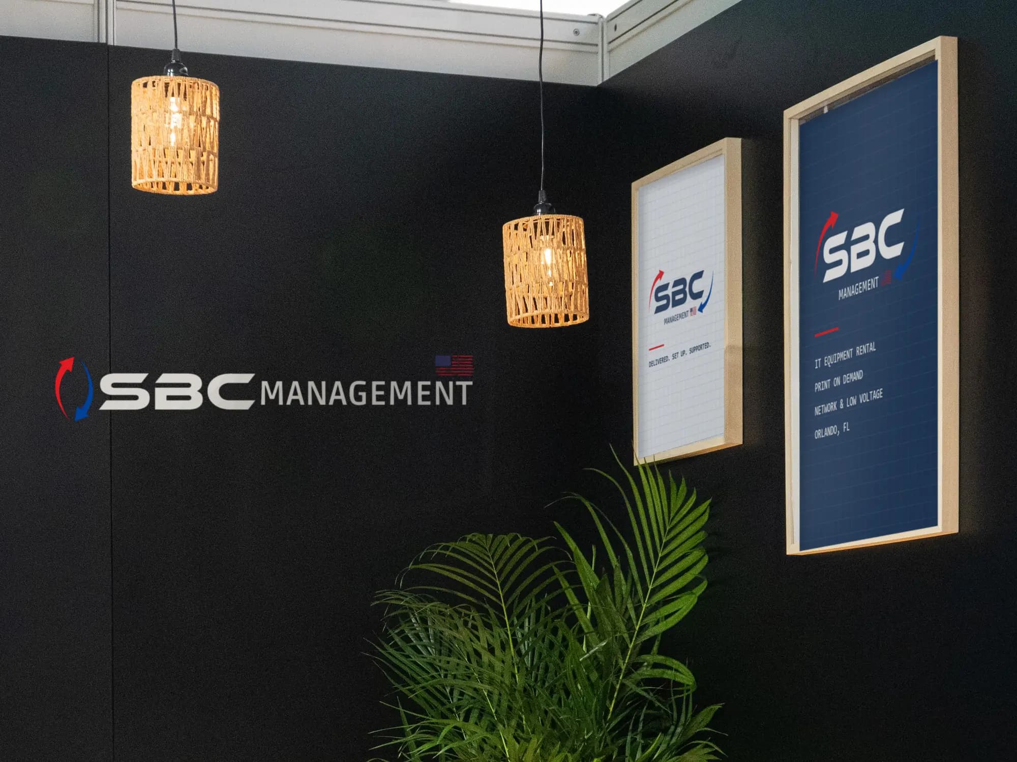 Trade show booth corner with the SBC Management logo on the wall, pendant lamps and framed SBC posters